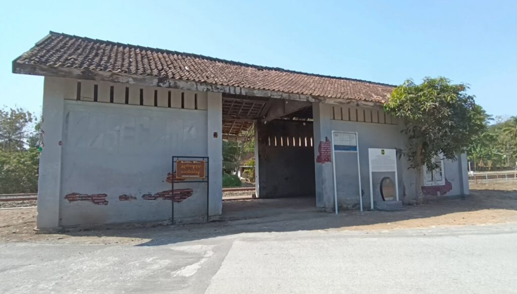 img-5335345 - Mabur.co Old small train station shelter with a tiled sloped roof, open central passage, and faded gray walls beside a tree.