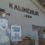 Cartoon man in overalls and cap smiling, holding a sign that reads 'Stasiun Lama' on a weathered wall with exposed bricks.