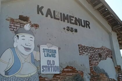 Cartoon man in overalls and cap smiling, holding a sign that reads 'Stasiun Lama' on a weathered wall with exposed bricks.