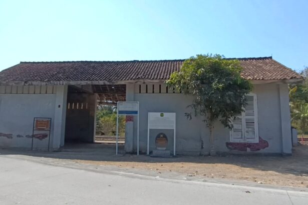 Small white concrete building with a red-tiled roof and an open central passage; a tree partly obscures the right side against a clear blue sky.