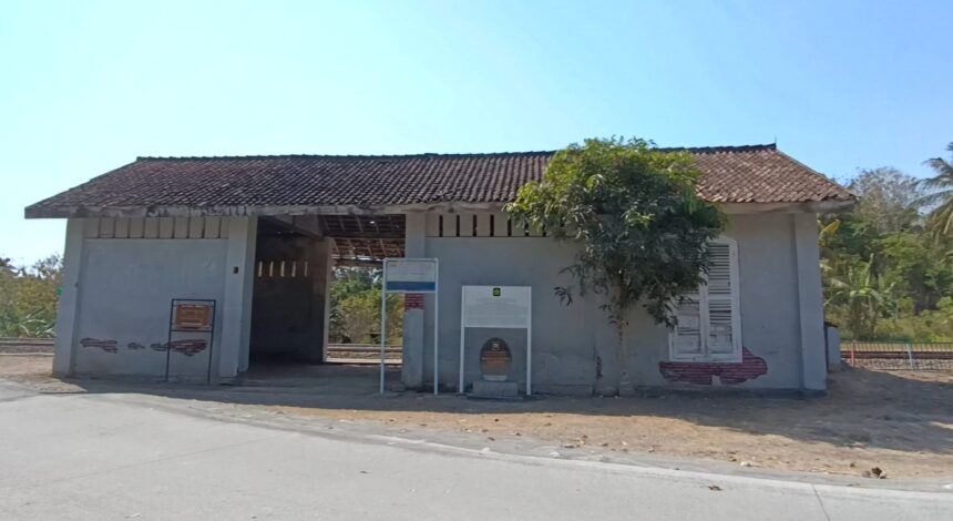 Small white concrete building with a red-tiled roof and an open central passage; a tree partly obscures the right side against a clear blue sky.