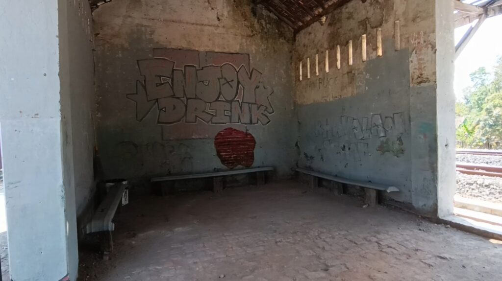 Abandoned railway shelter with graffiti on a worn wall and a red circular mark; benches along the wall and tracks outside visible to the right/entrance side.