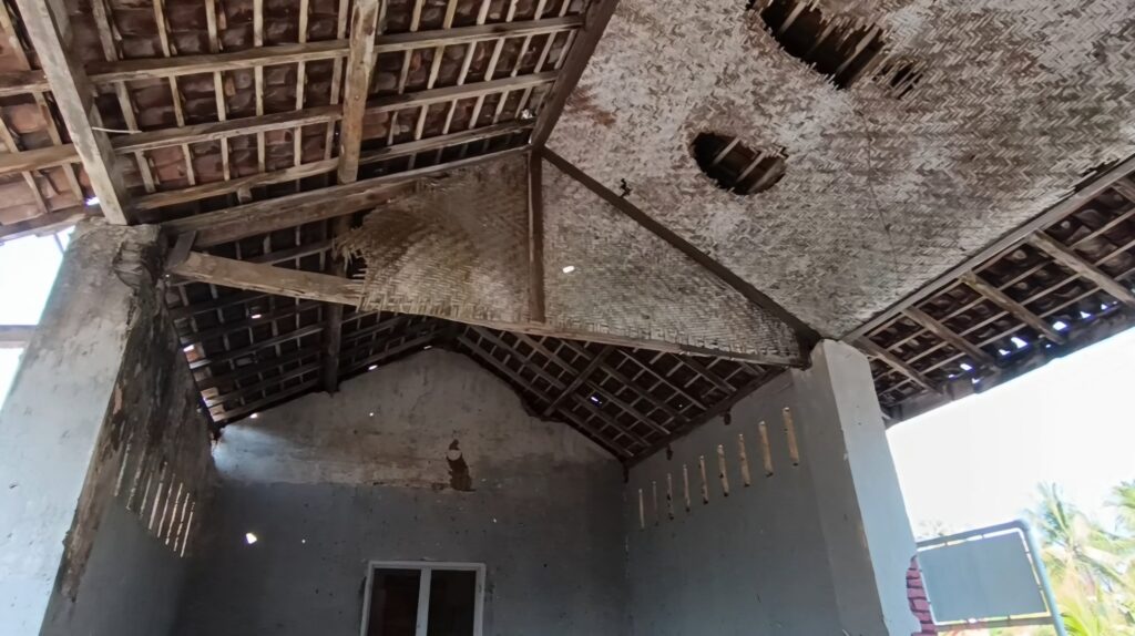 Interior of a dilapidated hut with a thatched roof, exposed wooden beams, and crumbling plaster walls with holes.