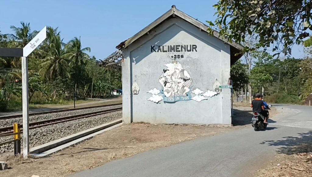 Small gray building by railway tracks with a mural reading 'KALIMENUR' and a person on a scooter on the road.