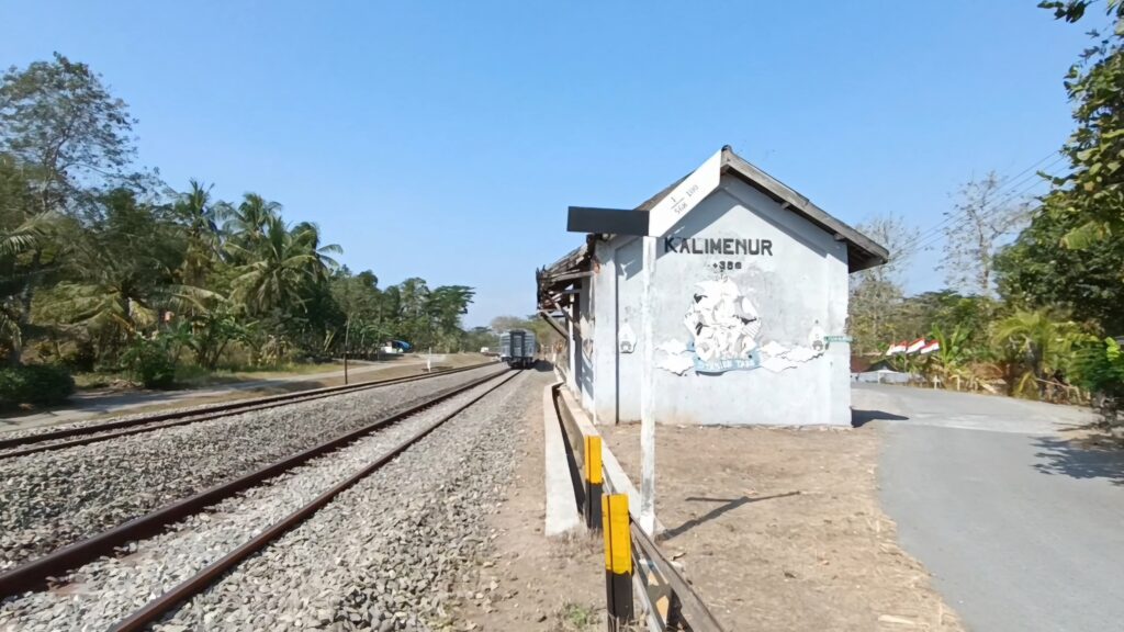 Rural railway platform with a small white station shed beside the tracks and trees under a clear blue sky.