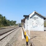Rural railway platform with a small white station shed beside the tracks and trees under a clear blue sky.