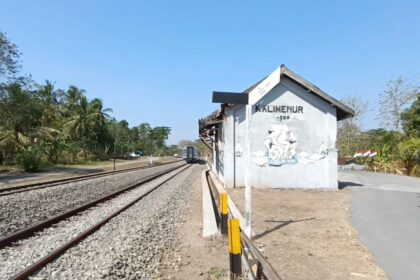 Rural railway platform with a small white station shed beside the tracks and trees under a clear blue sky.