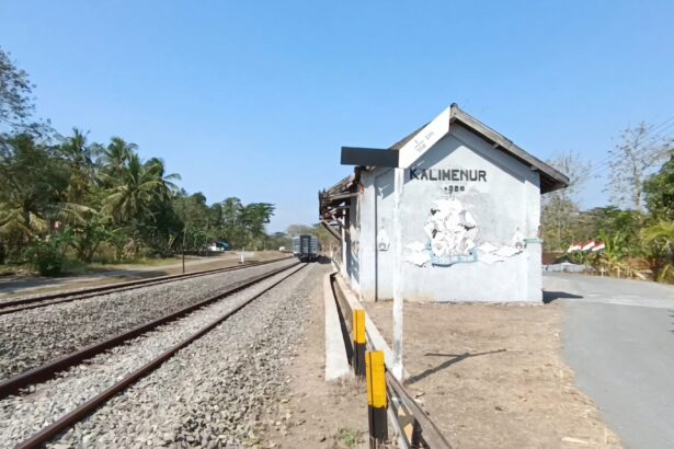 Rural railway platform with a small white station shed beside the tracks and trees under a clear blue sky.
