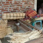 Woman sits on a bench weaving colorful palm leaf baskets outside a brick-walled workshop.