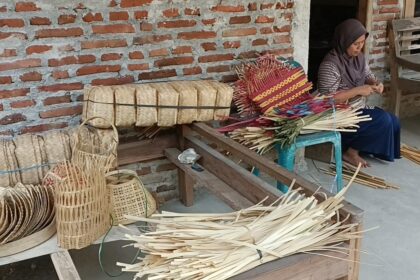 Woman sits on a bench weaving colorful palm leaf baskets outside a brick-walled workshop.