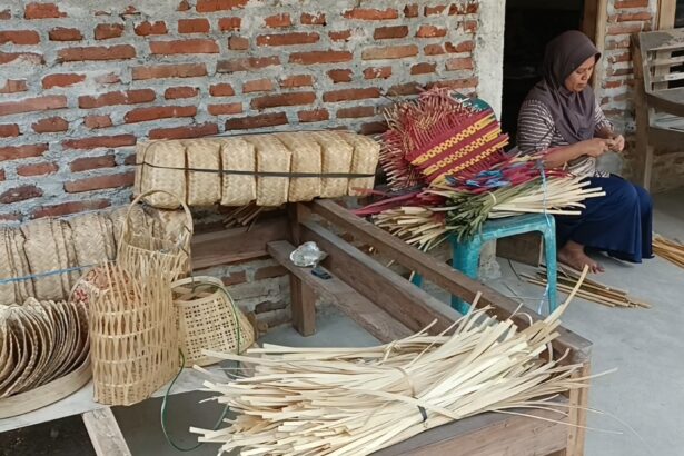 Woman sits on a bench weaving colorful palm leaf baskets outside a brick-walled workshop.