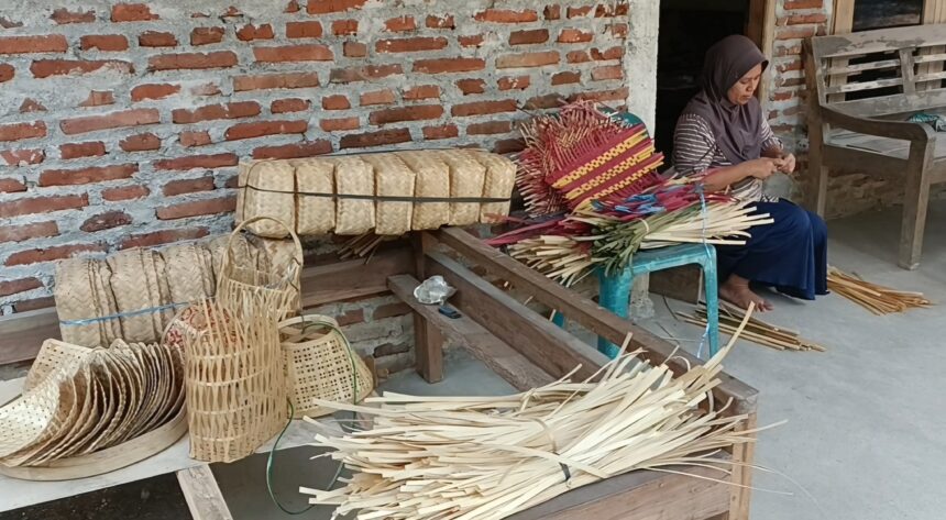 Woman sits on a bench weaving colorful palm leaf baskets outside a brick-walled workshop.