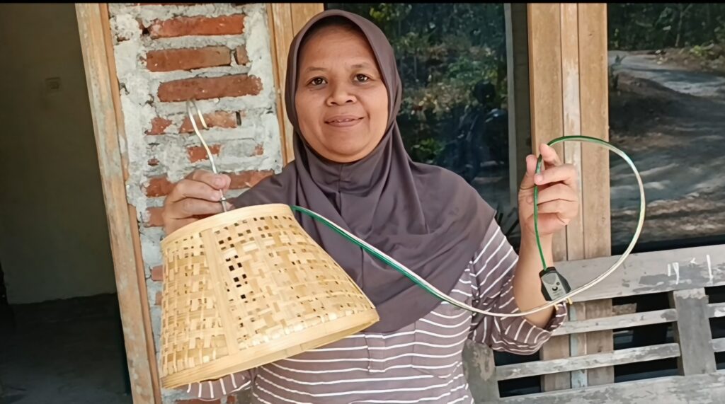 Smiling woman in a brown hijab and striped shirt holds a woven basket and a green hose outdoors beside a brick wall and wooden frame.
