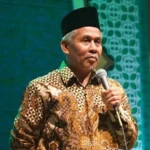 Man wearing a batik shirt and a black cap speaks into a microphone on stage.