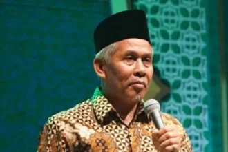 Man wearing a batik shirt and a black cap speaks into a microphone on stage.