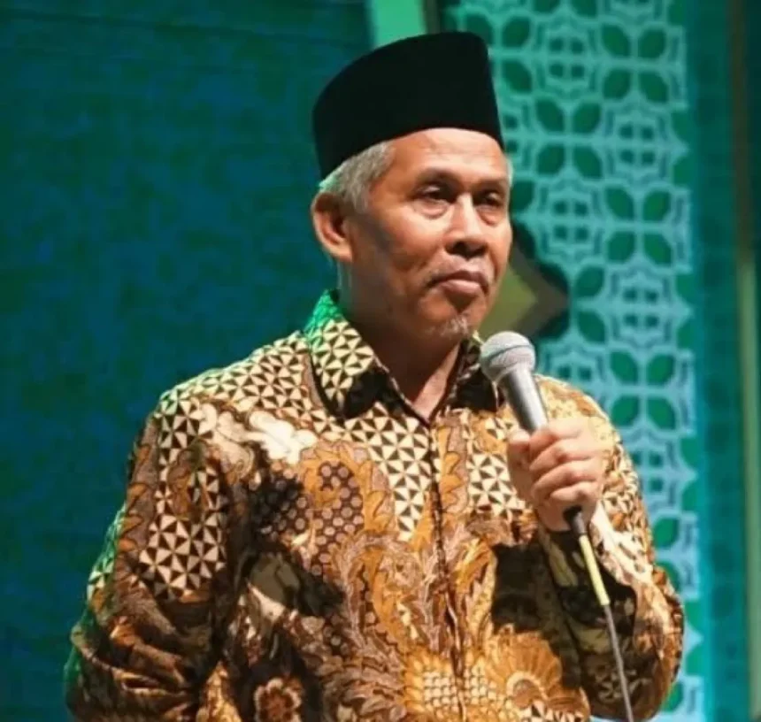 Man wearing a batik shirt and a black cap speaks into a microphone on stage.