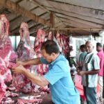 Butcher in a blue shirt carving a hanging beef carcass at an open-air meat market, with customers waiting nearby.