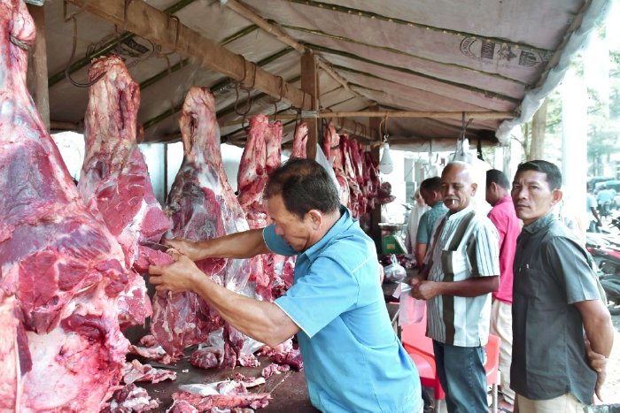 img_20240312_wa00081 - Mabur.co Butcher in a blue shirt carving a hanging beef carcass at an open-air meat market, with customers waiting nearby.