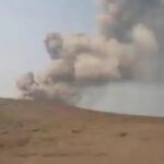 Desert landscape with a large plume of smoke rising from an explosion in the distance.