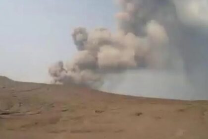 Desert landscape with a large plume of smoke rising from an explosion in the distance.