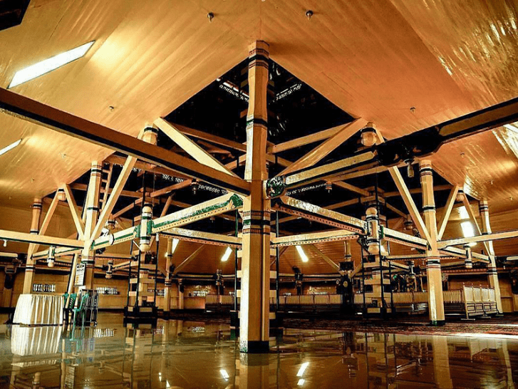 interior-masjid-agung-pondok-tinggi - Mabur.co Interior of a large timber-framed hall with a central pillar and crisscross support beams, warm lighting, and a glossy reflective floor.