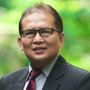 ioo - Mabur.co Portrait of a man wearing glasses, a dark suit, light shirt, and maroon tie, outdoors with a green, blurry background.