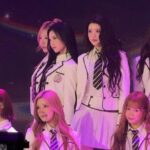 Six young women in white school-uniform outfits with striped ties pose on stage under purple lighting during a performance or photo op, some seated and others standing in two rows.