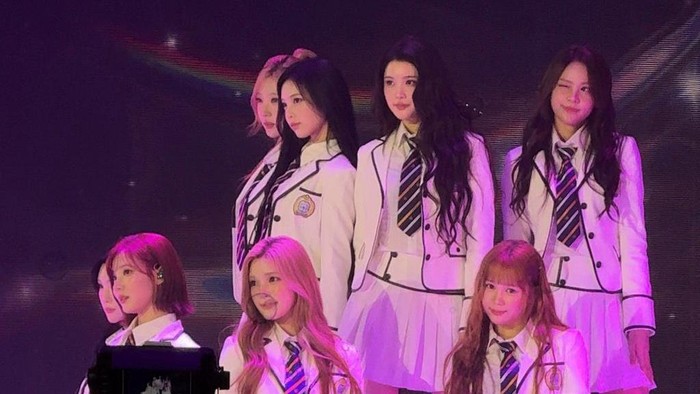 Six young women in white school-uniform outfits with striped ties pose on stage under purple lighting during a performance or photo op, some seated and others standing in two rows.