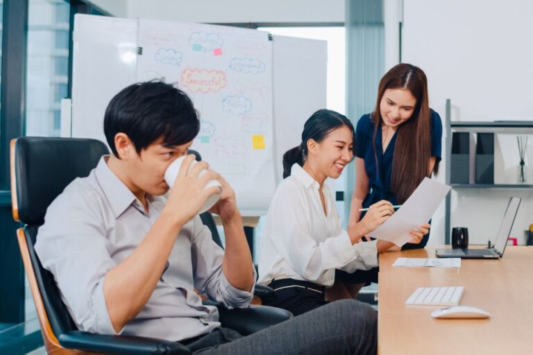 Three coworkers in an office: a man drinks from a cup while two women review a document at a shared desk, with a whiteboard in the background.
