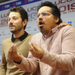 Two men at a press event gesturing as they speak, with sponsor banners in the background.