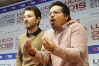 Two men at a press event gesturing as they speak, with sponsor banners in the background.