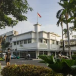 White multi-story building with the Indonesian flag on a central tower, framed by palm trees and greenery along a city street.