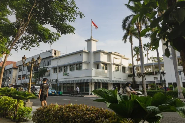 White multi-story building with the Indonesian flag on a central tower, framed by palm trees and greenery along a city street.