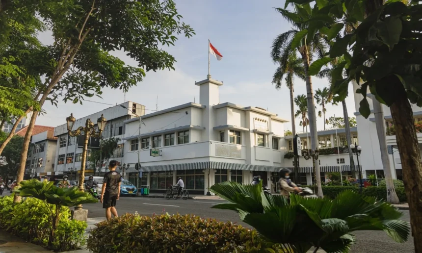 White multi-story building with the Indonesian flag on a central tower, framed by palm trees and greenery along a city street.