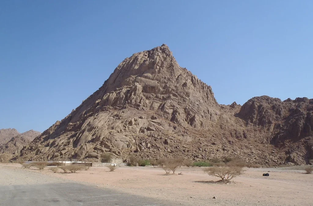 Desert landscape with a large jagged rocky hill under a clear blue sky; sparse trees and a small cluster of buildings at the base of the hill.