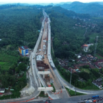 Aerial view of a long elevated highway under construction threading through a forested valley, with cranes, trucks, and a curved road below.