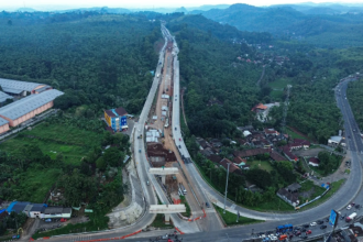 Aerial view of a long elevated highway under construction threading through a forested valley, with cranes, trucks, and a curved road below.