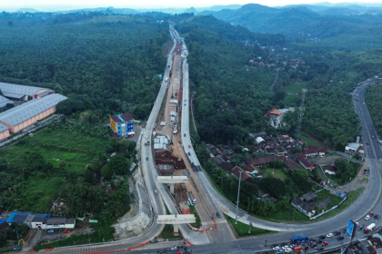 Aerial view of a long elevated highway under construction threading through a forested valley, with cranes, trucks, and a curved road below.