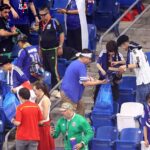 Group of fans in blue jerseys gathering in stadium seats, some holding bags and drinks.