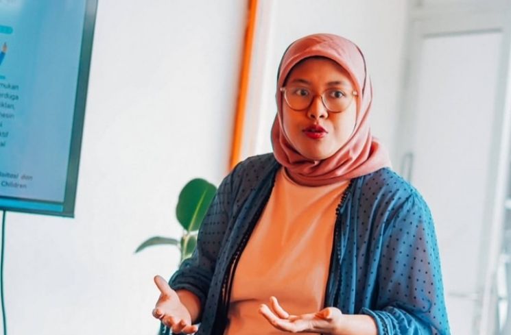 Woman in a pink hijab and glasses speaking while presenting, with hands gesturing near a monitor in a bright room.