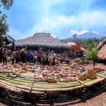 People browse a circular display of woven baskets and pottery around a bamboo platform under thatched huts in a village market setting.