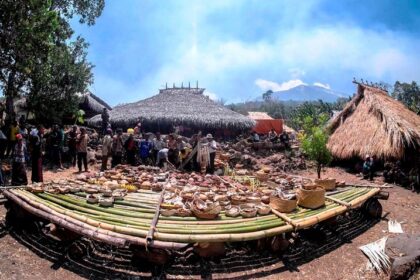 People browse a circular display of woven baskets and pottery around a bamboo platform under thatched huts in a village market setting.
