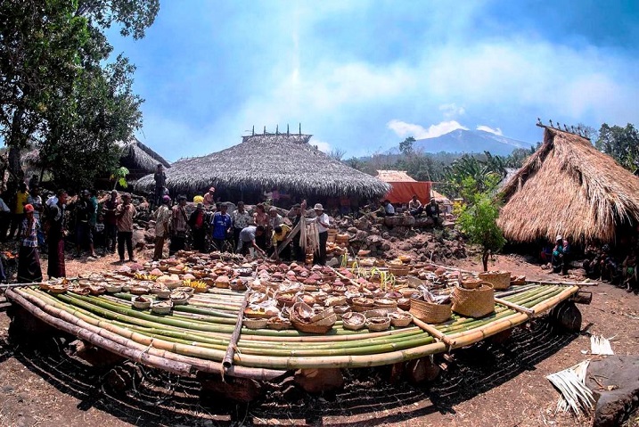 People browse a circular display of woven baskets and pottery around a bamboo platform under thatched huts in a village market setting.