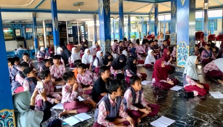 A large group of schoolchildren sits on the floor in a blue, decorated hall, listening and taking notes during a class or assembly.