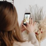 Person with brown hair photographing a dried floral arrangement with a smartphone in hand on a light interior setting.