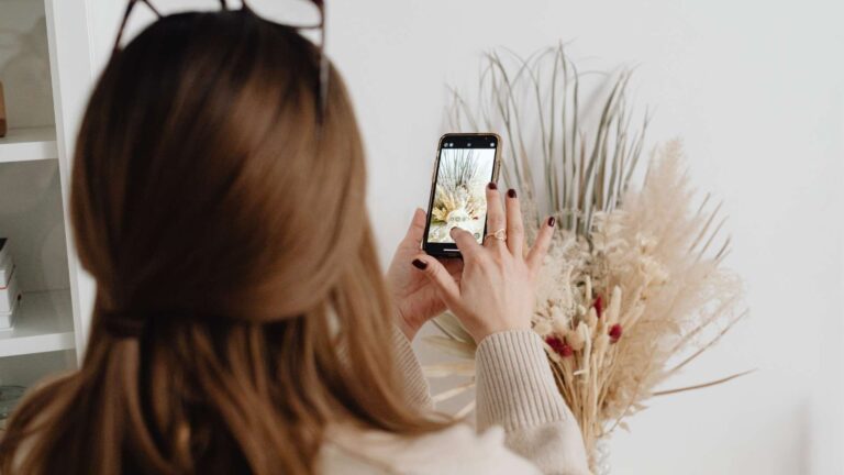 Person with brown hair photographing a dried floral arrangement with a smartphone in hand on a light interior setting.