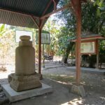 Outdoor shrine with a weathered stone monument under a corrugated metal roof, framed information boards nearby, and trees casting shade.