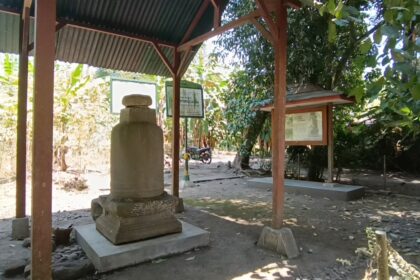 Outdoor shrine with a weathered stone monument under a corrugated metal roof, framed information boards nearby, and trees casting shade.