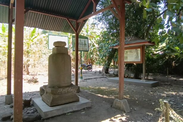 Outdoor shrine with a weathered stone monument under a corrugated metal roof, framed information boards nearby, and trees casting shade.