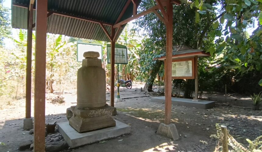 jjjh1 - Mabur.co Outdoor shrine with a weathered stone monument under a corrugated metal roof, framed information boards nearby, and trees casting shade.